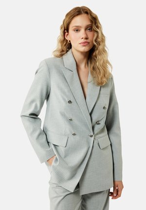 Junge Frau mit welligem blondem Haar, die einen hellgrauen, doppelreihigen Anzug trägt, mit den Händen in den Taschen vor schlichtem Hintergrund.