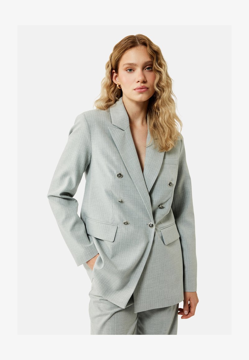 Ung kvinde med bølget blondt hår iført en lysegrå dobbeltradet dragt med hænderne i lommerne mod en ensfarvet baggrund.