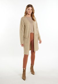 Cárdigan beige con bolsillos sobre una blusa beige y pantalones en color óxido. La modelo lleva botas de tobillo marrones y grandes pendientes coloridos.