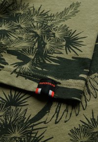 Oliven grøn T-shirt med mørkegrønne bladtryk. Har hvid syning og et sort mærke med røde og hvide detaljer. Blød bomuldsstruktur.