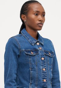 Denim jakna v srednje modri barvi z dvema prsnikoma, bakrenimi gumbi in klasičnim ovratnikom. Ima oprijet kroj in zapiranje na gumb.