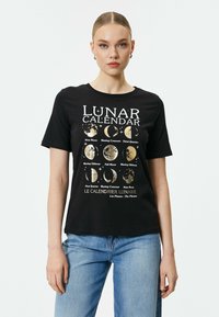 Sort bomulds t-shirt med et lunarkalender grafik i hvid og guld, med månefase illustrationer og tekst på flere sprog.