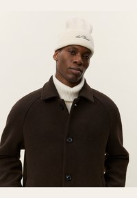 Brown wool coat with large black buttons and a soft cream knitted turtleneck. Features a light beige ribbed beanie with text detail.