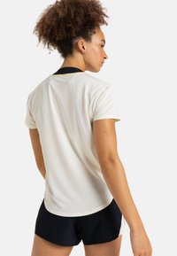 Camisa atlética de manga corta en un color crema claro con cuello negro. Presenta un corte relajado y acentos amarillos en los laterales.
