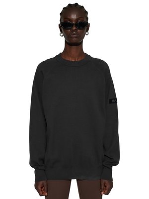 Vrouw met gevlochten haar draagt een zwarte oversized trui, donkere zonnebril, zilveren oorringen en een bruine broek tegen een witte achtergrond.