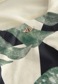 Green and black patterned fabric featuring a gold-toned clasp. The material is soft, with various leaf shapes and textures on a cream background.