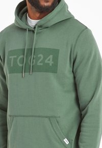 Olive green hoodie made of cotton blend, featuring a front kangaroo pocket, drawstring hood, and a large TON24 logo in a lighter hue.