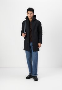 Svart hetteparkas med glidelås, brukt over en brun genser. Kombinert med løse blå jeans og svart fottøy, har ryggsekken en skulderrem.