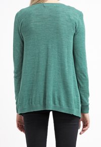 Vihreä pitkähihainen neulepaita pyöreällä kaula-aukolla, hienovaraisesti teksturoitua kangasta ja hieman pidempi takalaskos, yhdistettynä mustiin housuihin.