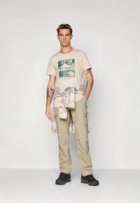 Bēša īsroku T-krekls ar zaļu grafisko zīmējumu, kombinēts ar gaismas krāsas cargo biksēm un melnām sporta kurpēm. Ap vidukli sasiets rakstains krekls.