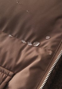 Braun wasserabweisender Stoff mit sichtbaren Tropfen, strukturiertem Oberflächen und einem Reißverschlussdetail am Rand, das seine Haltbarkeit und sein Design zur Schau stellt.