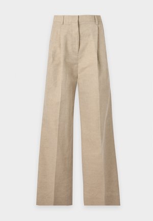 Femme debout portant un polo noir transparent à manches longues, un pantalon large beige et des chaussures noires pointues sur un fond clair.