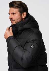 Chaqueta de plumas negra con cuello alto y capucha ajustable. Presenta una superficie texturizada y un parche con el logo en la manga izquierda. Material liso y aislante.