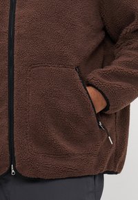 Personne portant une veste en polaire marron avec une fermeture éclair noire et une poche latérale, la main partiellement glissée dans la poche zippée.