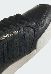 Černé tenisky Adidas s texturovanou kůží a semišovými panely, černými tkaničkami, zlatým logem Adidas a bílou pryžovou podrážkou.