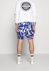 Blå och vita abstrakt mönstrade badshorts med elastisk midja, kombinerade med en vit tröja som har en cirkulär logotyp på baksidan.