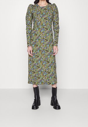 Femme portant une robe midi à manches longues avec épaules bouffantes et un imprimé floral vert, bleu et violet, associée à des bottines noires à talons.