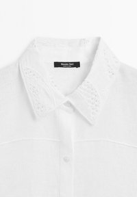 Chemise en lin blanc avec un col brodé, fermeture à boutons sur le devant et texture subtile. L'étiquette indique "Massimo Dutti."