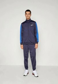 Navy sporta kostīms ar ziliem akcentiem, ar rāvējslēdzēju, ribotām aprocēm, logotipu uz krūtīm un kājām, kopā ar baltām sporta apavām neitrālā fonā.