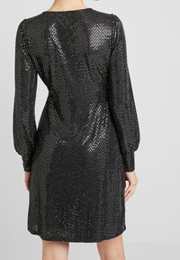 Černé šaty s přiléhavým střihem, dlouhými rukávy a texturovaným povrchem, který obsahuje rozptýlené flitry pro přidaný třpyt a vzor.