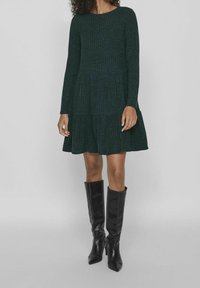 Robe côtelée vert foncé à manches longues, décolleté rond et jupe évasée, associée à des bottes noires à talons hauts arrivant au genou.