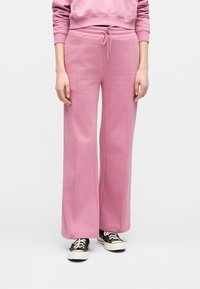 Άτομο που φοράει ροζ φαρδιά φόρμα με κορδόνι στη μέση και μαύρα αθλητικά χαμηλού τύπου, μπροστά σε απλό φόντο.