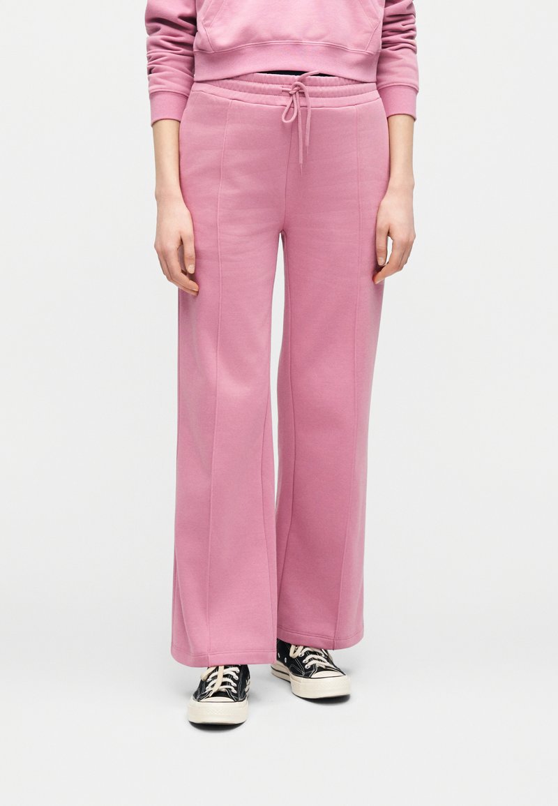 Άτομο που φοράει ροζ φαρδιά φόρμα με κορδόνι στη μέση και μαύρα αθλητικά χαμηλού τύπου, μπροστά σε απλό φόντο.