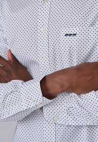 Chemise blanche à manches longues avec un petit motif en losanges bleu marine, col boutonné et une poche poitrine ornée de trois figures brodées en bleu marine.