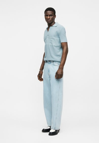 Άνδρας που στέκεται με ανοιχτό γαλάζιο πόλο πουκάμισο, χαλαρά τζιν ανοιχτού ξεβαμένου χρώματος και ασπρόμαυρα επίσημα παπούτσια, μπροστά σε ένα απλό λευκό φόντο.