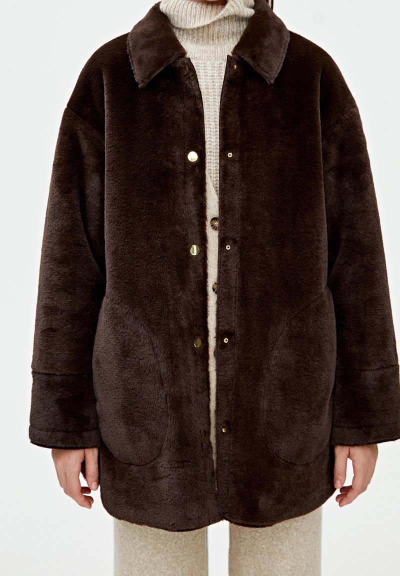 Brown fleece jacket with a collar, snap buttons, and side pockets. The design features a soft texture and relaxed fit, layered over a beige sweater.