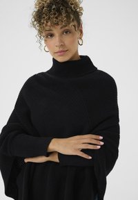 Czarny dzianinowy sweter z golfem o luźnym kroju, z prążkowaną fakturą i szerokimi rękawami. Model pozuje z założonymi rękami.