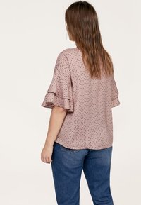 Lyserød blouse med prikket mønster og flæser på ærmerne; har runde prikker i sort og hvid, letvægtsstof og løstsiddende pasform. Bæres med blå jeans.