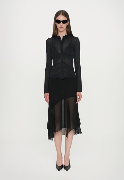 Black sheer long-sleeve top with button closure and gathered design paired with a black asymmetrical hem skirt. Black pointed-toe heels.