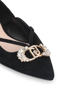 Chaussure noire en daim à bout pointu avec une boucle dorée ornée de cristaux et un design à croisillons. Fini intérieur lisse.