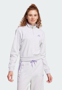 Sivi atletski pulover s visokim ovratnikom, zatvaranjem na zip i elastičnim pojasom. Ima ljubičaste akcenta i logo na prsima i hlačama.