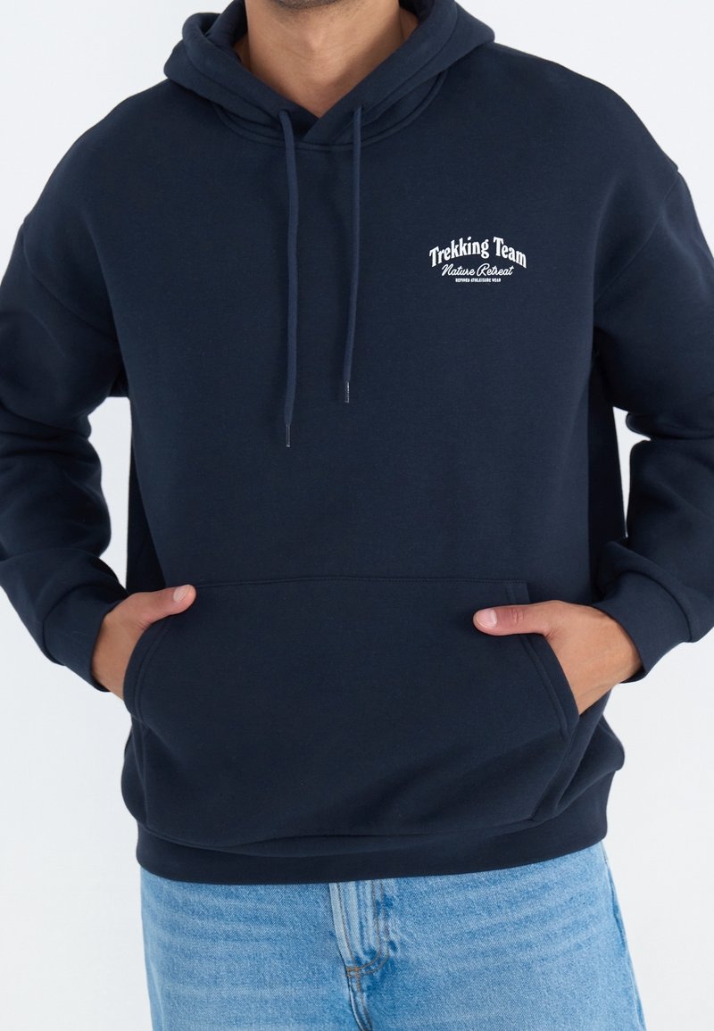 Homme portant un sweat à capuche bleu marine avec poche avant et texte « Trekking Team Nature Retreat » sur la poitrine, associé à un jean bleu clair.