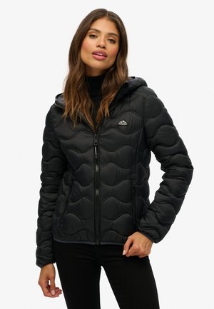 Vrouw in een zwarte gewatteerde hooded Jack met rits en logo, poserend met één hand bij haar taille op een witte achtergrond.