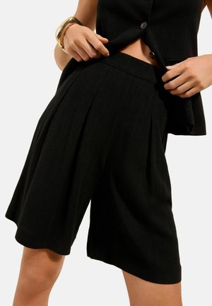 Mujer con pantalones cortos negros de pierna ancha hasta la rodilla y un top sin mangas a juego con botones, ajustándose el dobladillo del top, con una pulsera dorada en la muñeca.