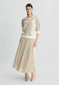 Felpa dorata con paillettes, scollo rotondo e maniche lunghe, abbinata a una lunga gonna plissettata coordinata. Tacchi classici a punta.