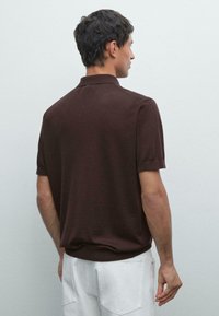Polo shirt a maglia marrone con maniche corte e colletto classico, caratterizzato da un orlo a coste e una texture liscia. Indossato con pantaloni bianchi.