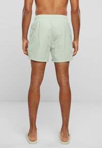 Lysegrøn badetights med elastisk talje, laget av teksturert stoff og med side lommer. Kort lengde, brukt med beige sandaler.