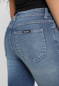 Jeans de mezclilla azul con un diseño ajustado, que presentan una costura de bolsillo curva distintiva y una etiqueta en el bolsillo trasero. Textura ligeramente desgastada.