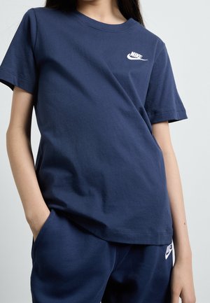 Person iført en mørkeblå Nike t-shirt og matchende joggingbukser, med den ene hånd i lommen, foran en ensfarvet lys baggrund.