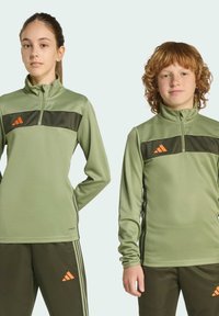Zelené športové zip-top tričká s čiernymi akcentmi, s logom Adidas v oranžovej farbe, v kombinácii so zelenými nohavicami so pruhmi po stranách.