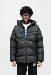 Czarna kurtka puchowa z kapturem, ozdobiona pikowanym wzorem, z przednim zamkiem błyskawicznym i kieszeniami po bokach, noszona na białej koszuli i ciemnych spodniach.