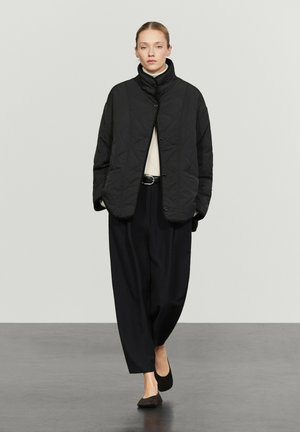 Frau trägt eine schwarze gesteppte Jacke, lockere schwarze Hose, beige Rollkragenpullover und schwarze Ballerinas, steht vor einem schlichten hellen Hintergrund.