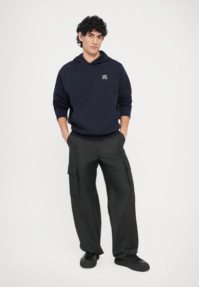 Σκούρα μπλε φούτερ με κουκούλα και λογότυπο στο μπροστινό μέρος, συνδυασμένο με φαρδιά μαύρα παντελόνια cargo με πλαϊνές τσέπες και μαύρα αθλητικά παπούτσια.