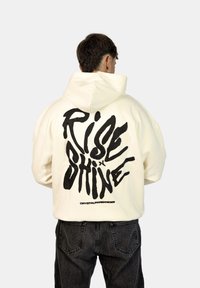 Felpa con cappuccio color crema con una grande scritta grafica nera "RISE x SHINE" sul retro, dal taglio rilassato e dalla texture morbida.