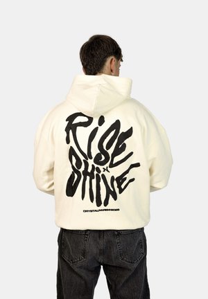 Crème-kleurige hoodie met een groot zwart grafisch tekst "RISE x SHINE" op de achterkant, met een relaxte pasvorm en een zachte textuur.