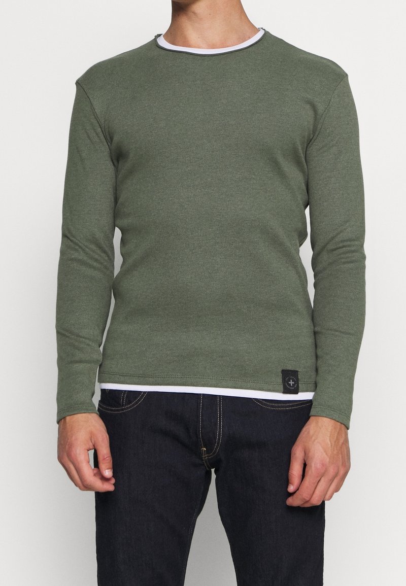 Homme portant un t-shirt vert olive à manches longues et col rond par-dessus un sous-vêtement blanc, associé à un jean bleu foncé sur un fond uni.